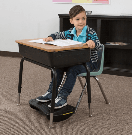 Menino sentado em carteira escolar apoiando os pés em um apoio de pé preto